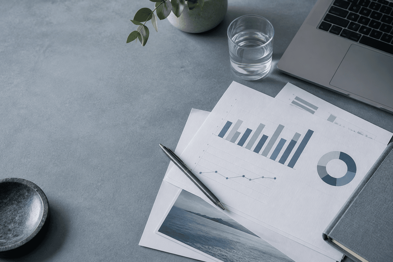 Printed marketing charts, a pen, a glass and a laptop arranged on a cool blue-grey workspace